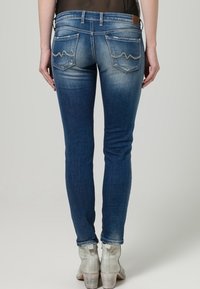 Jeans ajustados de denim azul oscuro con acentos descoloridos, que presentan bolsillos cosidos con un diseño curvado, combinados con botas de tobillo claras.