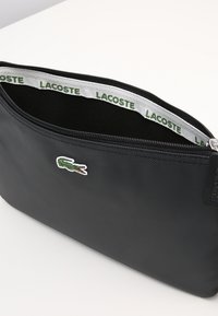 Pochette nera in nylon con chiusura a zip, dotata di un piccolo logo bianco Lacoste e di una fodera interna verde marchiata. Superficie texture.