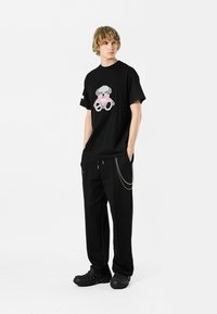 Camiseta negra de manga corta con un gráfico de oso de peluche colorido, combinada con pantalones negros y calzado negro texturizado con un detalle de cadena.