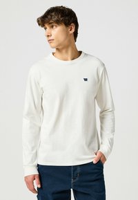 Biała, bawełniana koszulka z długim rękawem, z okrąg łym dekoltem i małym granatowym logo po lewej stronie na klatce piersiowej, zestawiona z ciemnoniebieskimi dżinsami.