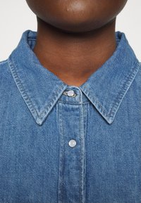 Camicia in denim con colletto, di colore azzurro chiaro, dotata di chiusura con bottoni al centro e dettagli di cucitura visibili sul tessuto.
