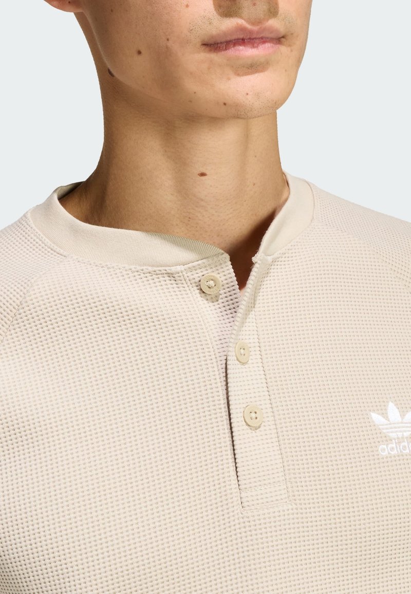 Beige, textured henley shirt with a round collar and three buttons. Features a small white logo on the left chest.