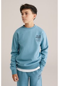 Svetlo modra majica z dolgimi rokavi iz mehkega materiala, z rebrastimi manšetami in ovratnikom. Na sprednji strani je napisana besedilo "BLUE RIDGE ATHLETIC" v temnem tisku.