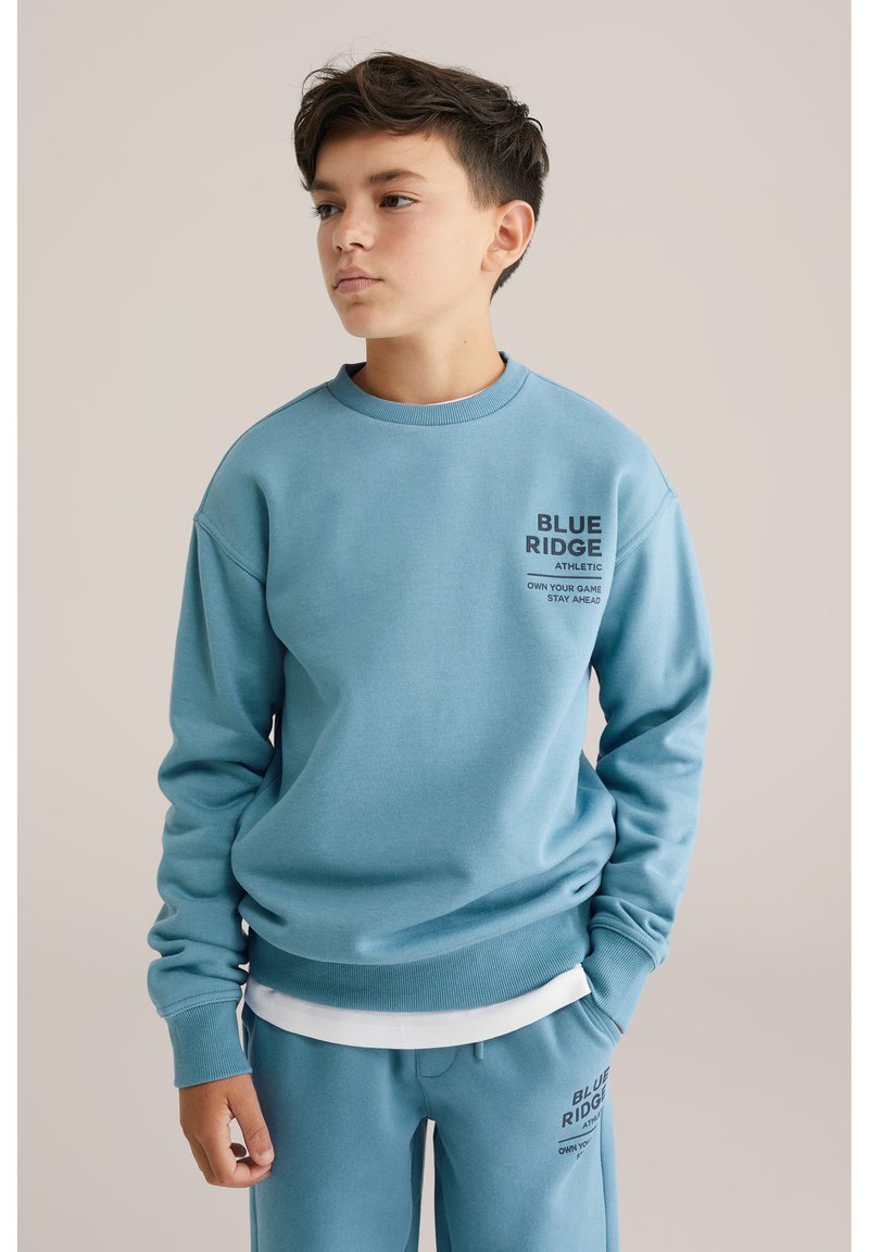 Svetlo modra majica z dolgimi rokavi iz mehkega materiala, z rebrastimi manšetami in ovratnikom. Na sprednji strani je napisana besedilo "BLUE RIDGE ATHLETIC" v temnem tisku.
