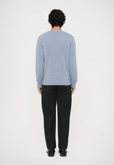 Mężczyzna z kręconymi włosami, ubrany w jasnoniebieski sweter, czarne tapered pants i czarne buty, stojący tyłem na białym tle.