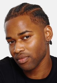 Gros plan sur une personne avec des cheveux tressés, une peau lisse, une boucle d'oreille discrète et une expression neutre, portant un shirt noir. Fond blanc.