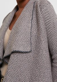 Kabát s houndstooth vzorem v odstínech šedé a béžové, který se vyznačuje jemnou texturou, otevřeným předním dílem a designem s šálovým límcem.