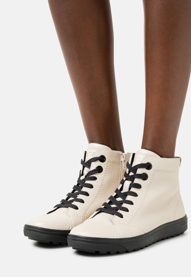 Paire de bottines beiges à lacets et semelles noires portées sur des jambes nues à peau foncée sur fond blanc.