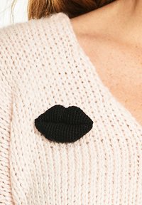 Czarny broszka w kształcie ust z dzianiny na jasnoróżowym swetrze o grubej dzianinie. Broszka ma trójwymiarowe, teksturowane detale.