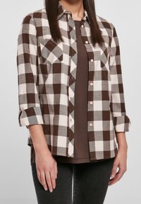 Femme portant une chemise à carreaux marron et blanc sur un t-shirt marron et un jean foncé, debout avec les bras détendus le long du corps.