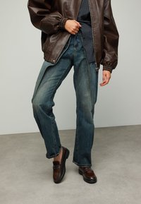 Bruine leren bomberjack over een blauw geruit overhemd, gecombineerd met wijde blauwe spijkerbroeken en donkerbruine instapschoenen.