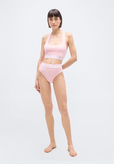 Γυναίκα που στέκεται ξυπόλητη φορώντας ανοιχτό ροζ σλιπ με ψηλή μέση και ασορτί αθλητικό σουτιέν, μπροστά σε λευκό φόντο.