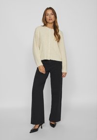Cardigan crema e morbido con scollatura rotonda e chiusura con bottoni, abbinato a pantaloni neri a gamba larga e tacchi neri. Tessuto liscio e strutturato.