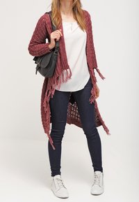Un long cardigan en tricot bordeaux à l'avant ouvert avec des bordures à franges, superposé à un haut blanc. Porté avec un jean foncé et des baskets blanches, tenant un sac à main.
