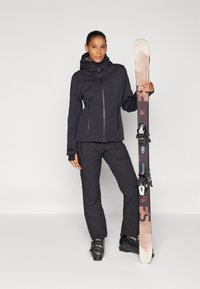 Zwarte skijas met ritssluiting, capuchon en twee voorzakken. Zwarte geïsoleerde skibroek en skischoenen. Model houdt een paar ski's met bindingen vast.