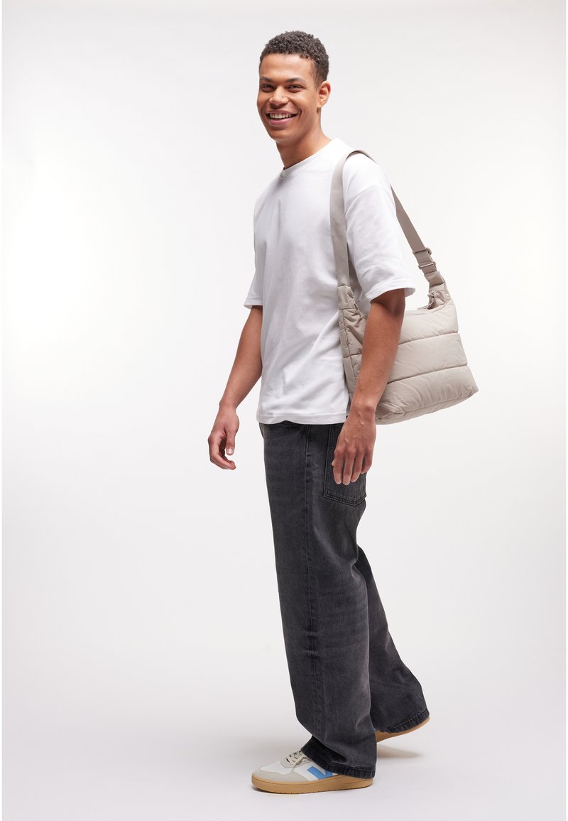 Joven sonriente, vestido con camiseta blanca, vaqueros negros, zapatillas y llevando un bolso beige acolchado al hombro sobre un fondo blanco liso.