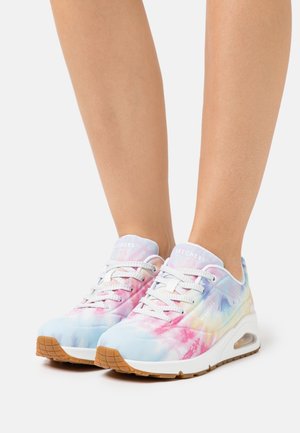 Personne portant des baskets à plateforme blanches avec des motifs tie-dye pastel en rose, bleu et jaune, avec des lacets blancs et des semelles marron.