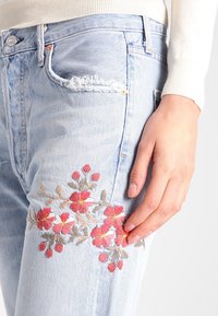 Světle modré džíny z denimu s opotřebovanou záplatou, zdobené složitou květinovou výšivkou v červené a béžové barvě na stehně, s přiléhavým střihem.