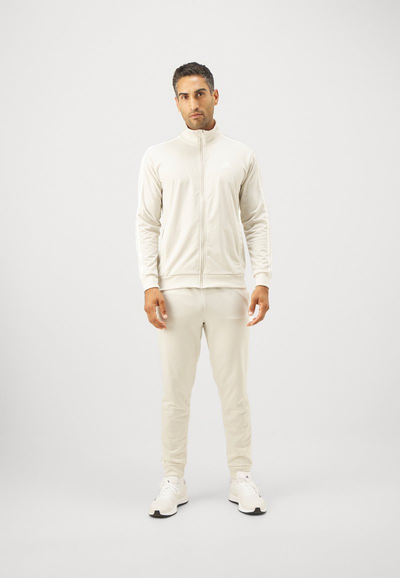 Svetlobežový teplákový komplet vyrobený z hladkej tkaniny. Sako na zips s vysokým golierom, zladené nohavice a biele športové topánky.