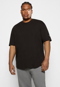 T-shirt in cotone nero con vestibilità comoda, maniche corte e un patch con logo sulla manica sinistra. Indossato con pantaloni della tuta grigi.