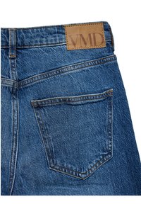 Poche arrière d'un jean en denim bleu avec des coutures visibles et un patch en cuir marron marqué VMD au-dessus de la poche.