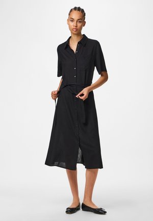 Kvinde justerer bælte på sort, kortærmet, midikjole med knapper foran og krave, kombineret med sorte balletsko, stående på hvid baggrund.