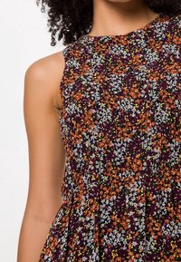 Robe sans manches avec un motif floral violet, orange, blanc et vert, portée par une personne aux cheveux bouclés et foncés, coupée au niveau des épaules et de la poitrine.