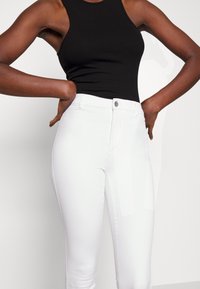 Top de malha ranhurada preta combinado com jeans brancos ajustados com fecho de botão e textura limpa e suave.