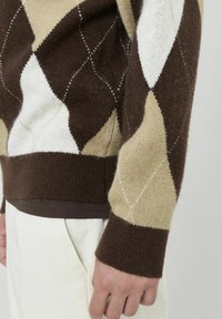 Pull en argyle marron avec des motifs de losanges crème et beige, fabriqué en matière tricotée douce. Il comporte des poignets et un ourlet à côtes, avec une chemise foncée superposée.