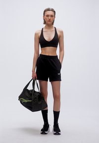 Γυναίκα με μαύρο αθλητικό σουτιέν, σορτς, κάλτσες και αθλητικά παπούτσια, κρατώντας μαύρη τσάντα γυμναστηρίου, στέκεται μπροστά σε απλό φωτεινό φόντο.