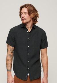 Camicia nera con maniche corte e bottoni in un tessuto testurizzato, dotata di colletto, leggero lucido e dettaglio con etichetta laterale.