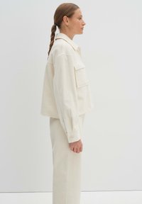 Cremefarbene Cordjacke mit einem cropped Design, ausgestattet mit einem Kragen und zwei Vorderseiten-Taschen, kombiniert mit passenden Hosen. Glatte Textur, gerader Schnitt.