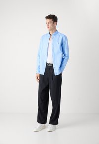 Camicia azzurro chiaro con maniche lunghe, indossata sopra un top bianco. Abbinata a pantaloni neri a righe e sneakers bianche.
