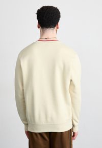 Sudadera de color beige claro, hecha de material suave, con cuello redondo y puños acanalados. La parte trasera presenta un mínimo de detalles y una textura lisa.