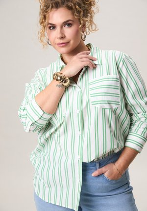 Kroeshaar vrouw met een groen-wit gestreept shirt en blauwe spijkerbroek, geaccentueerd met een gouden armband, oorringen en een kettinkje.