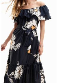 Sininen off-the-shoulder -mekko, jossa on röyhelökaulus, valkoisia kukkakuviota ja oranssit keskustat, vyötäröllä istuvalla yhteensopivalla vyöllä.