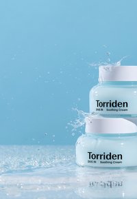 Kaksi läpikuultavaa purkkia Torriden Soothing Creamia vaaleansinisen värisenä valkoisilla kansilla, ympäröitynä vesiroiskeilla kiiltävällä pinnalla.