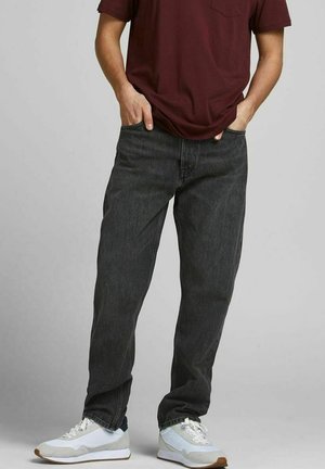 Person wearing dark gray jeans, white sneakers, and a burgundy t-shirt with hands in pockets against a plain gray background.