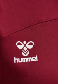 Camiseta deportiva burdeos con tejido texturizado. Presenta un logo blanco con un diseño estilizado de abeja y la palabra "hummel" debajo.