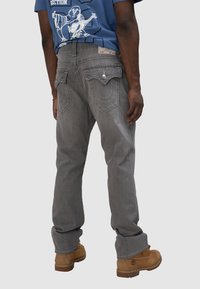 Jeans de mezclilla gris con un ajuste relajado, que cuentan con bolsillos traseros con costuras distintivas y una cintura etiquetada, combinados con botas de trabajo marrones.