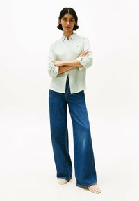 Camicia button-up a righe verde chiaro con maniche arrotolate, abbinata a pantaloni in denim blu a gamba larga e scarpe beige a punta.