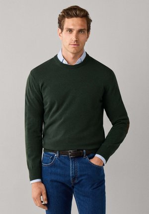 Homme portant un pull vert foncé par-dessus une chemise bleu clair à col et un jean bleu, debout avec une main dans la poche, sur un fond uni.