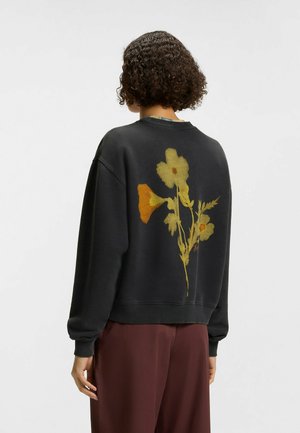 Persoon met kort krullend haar draagt een zwarte sweater met een geel-oranje bloemmotief op de achterkant, gecombineerd met losse bruine broek.