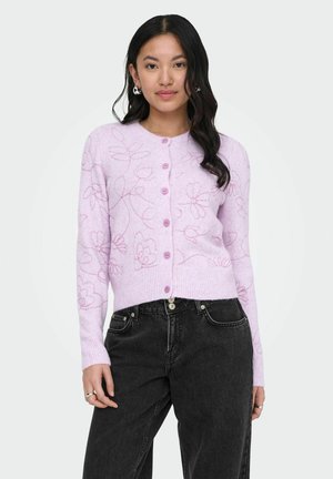 Žena oblečená v levanduľovom zapínacom kardigane s kvetinovými vzormi a čiernych džínsach s vysokým pásom, stojaca pred jednofarebným pozadím.