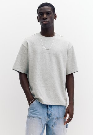 Jeune homme portant un T-shirt oversize gris clair, un jean en denim bleu clair, un collier en chaîne argentée, un bracelet et une bague, debout devant un fond uni.
