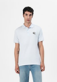 Lyseblå polo skjorte laget av bomull med en myk tekstur, med krage, tre knapper og et lite svart-hvitt grafisk trykk på brystet.