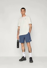 Sportska odjeća koja uključuje bijelu majicu, mornarsko plave kratke hlače s logotipom, crne tenisice i crnu bocu za vodu; kratke hlače imaju laganu teksturu.