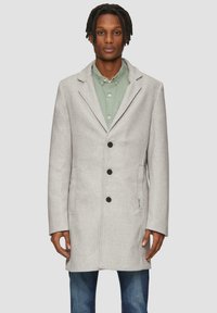 Grijze wollen mix jas met een geknipt kraag, eennelzijdig ontwerp, drie zwarte knopen en zijzakken, gedragen over een groene shirt.