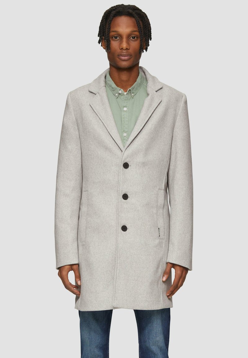 Grijze wollen mix jas met een geknipt kraag, eennelzijdig ontwerp, drie zwarte knopen en zijzakken, gedragen over een groene shirt.