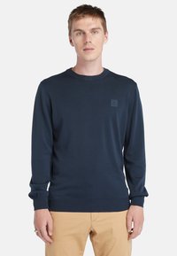 Marineblauwe gebreide trui met een geribbelde ronde halslijn, lange mouwen en een subtiele logopatch op de borst. Vergezeld door kaki broek.